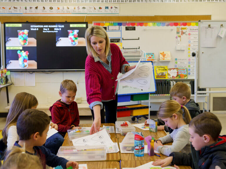 A teacher hands worksheets to a group of young students seated at a table.